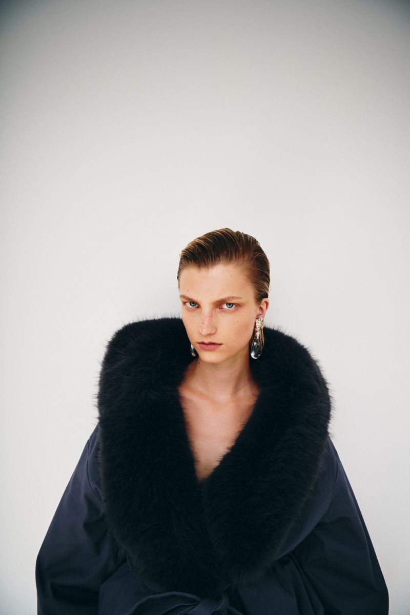 Model with slicked-back hair wearing a dark coat with a large fur collar against a white background