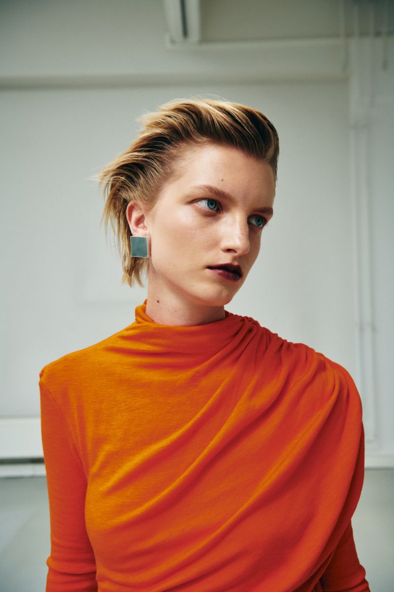 Model with slicked-back blonde hair wearing an orange turtleneck and square metallic earrings against a light studio backdrop.
