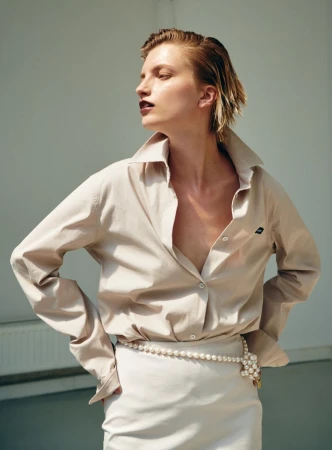 Model with slicked-back blonde hair wearing a beige button-down shirt and white skirt against a plain wall.