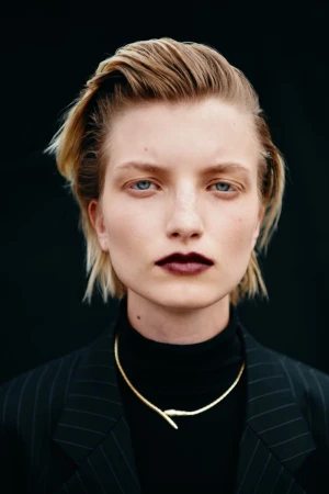 Close-up portrait of a woman with slicked-back blonde hair, dark lipstick, gold choker necklace, and a black pinstripe blazer.