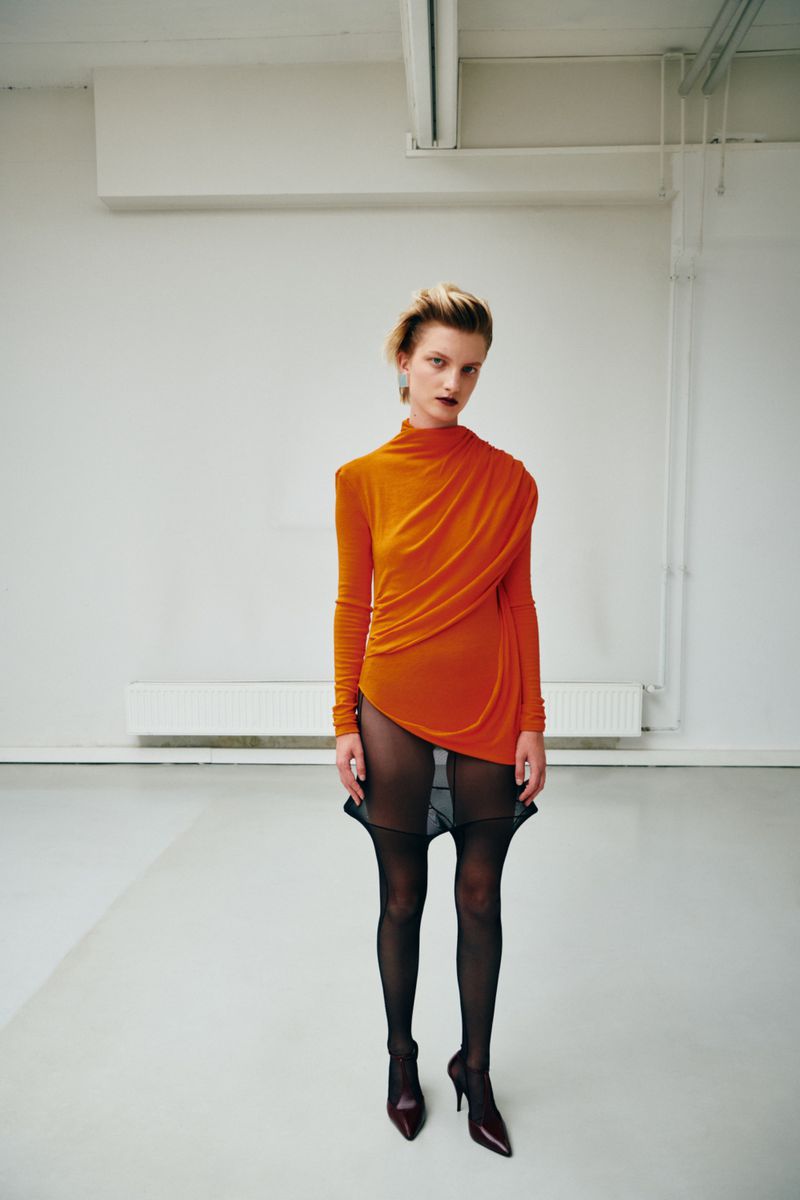 Model in an orange draped top and sheer black tights standing against a plain white interior wall