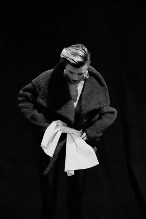 Model wearing a textured dark coat with a large white tied belt against a black background