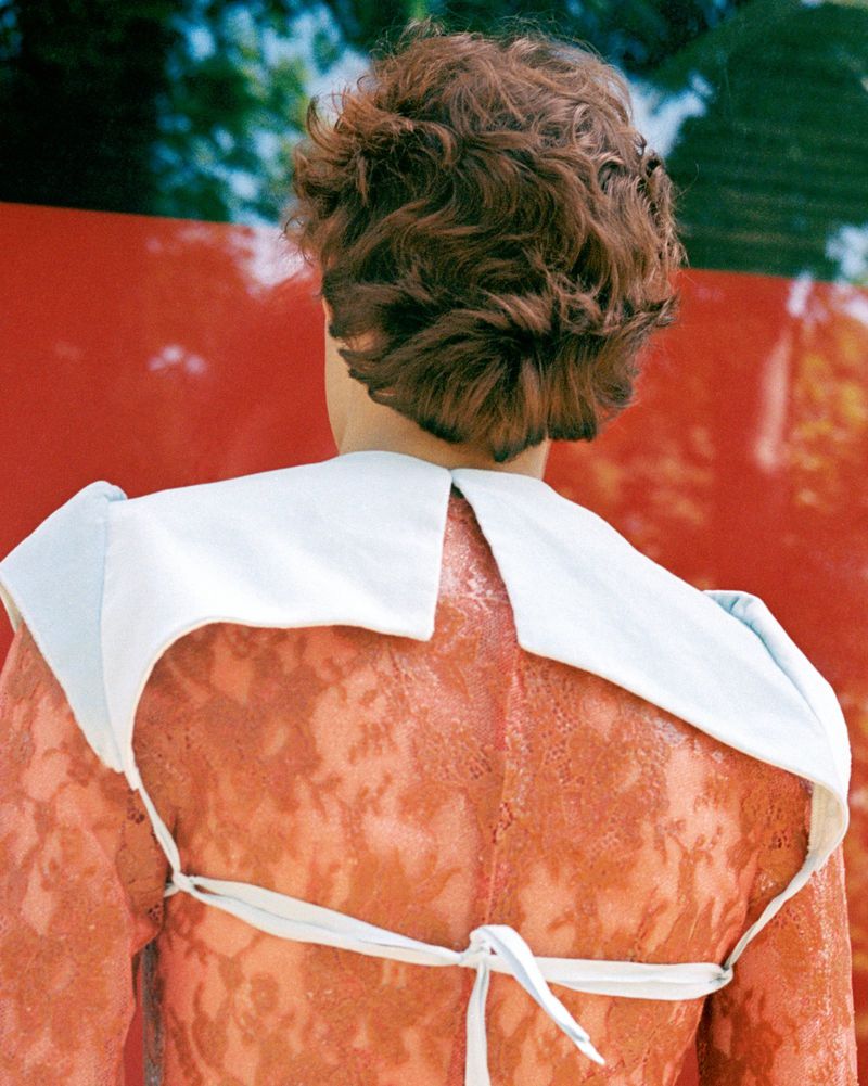 Rear view of a model with short curly brown hair wearing a sheer lace garment against a bright red background.