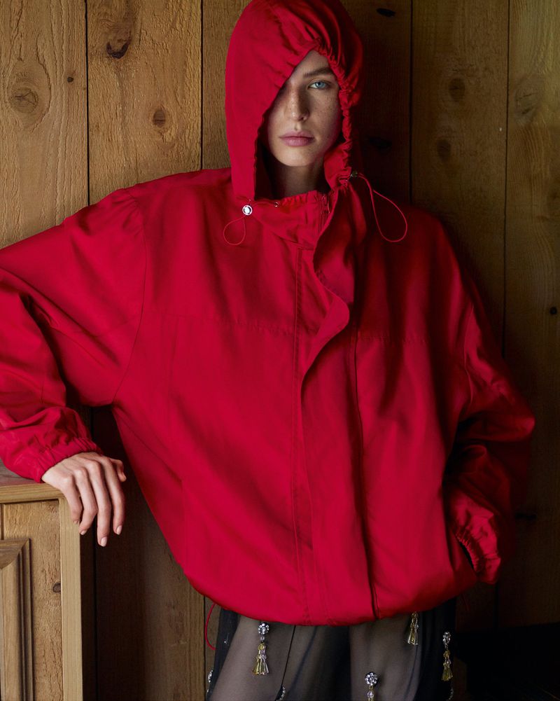 Model wearing a bright red hooded jacket standing against a vertical wood paneled wall.