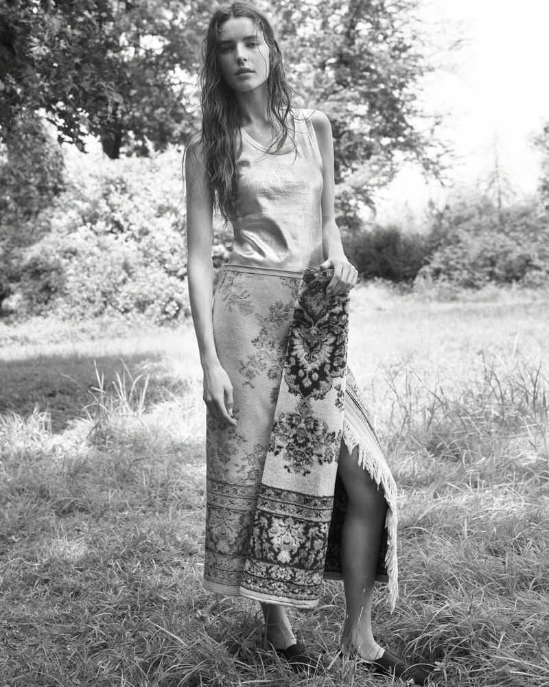 Model standing outdoors in a grassy field wearing a patterned skirt and holding a fringed textile for The Gentlewoman.