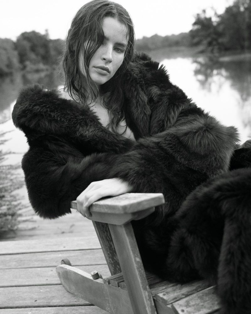 Black and white portrait of a woman in a dark fur coat sitting on a wooden chair by a lake.