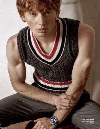 Close-up portrait of a red-haired male model wearing a sleeveless grey knit sweater vest with red and white trim.