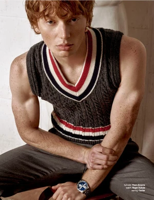 Close-up portrait of a red-haired male model wearing a sleeveless grey knit sweater vest with red and white trim.