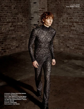 Model Calum Harper wearing a dark metallic patterned jumpsuit in an industrial brick room