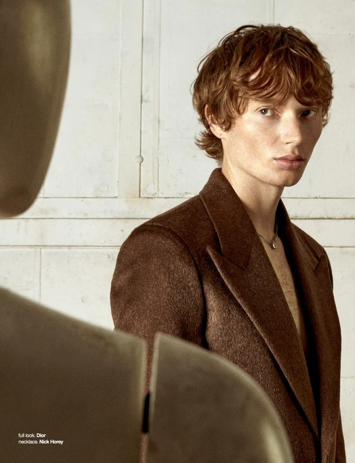 A young male model with reddish-brown hair wearing a brown blazer against a textured white wall