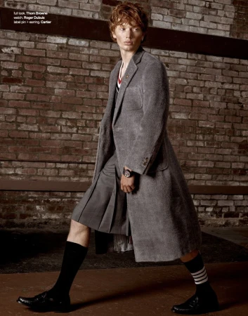 Model with red hair wearing a grey coat and pleated skirt against a weathered brick wall