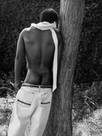 Shirtless man with a white fabric draped around his neck leaning against a tree in a black and white editorial portrait