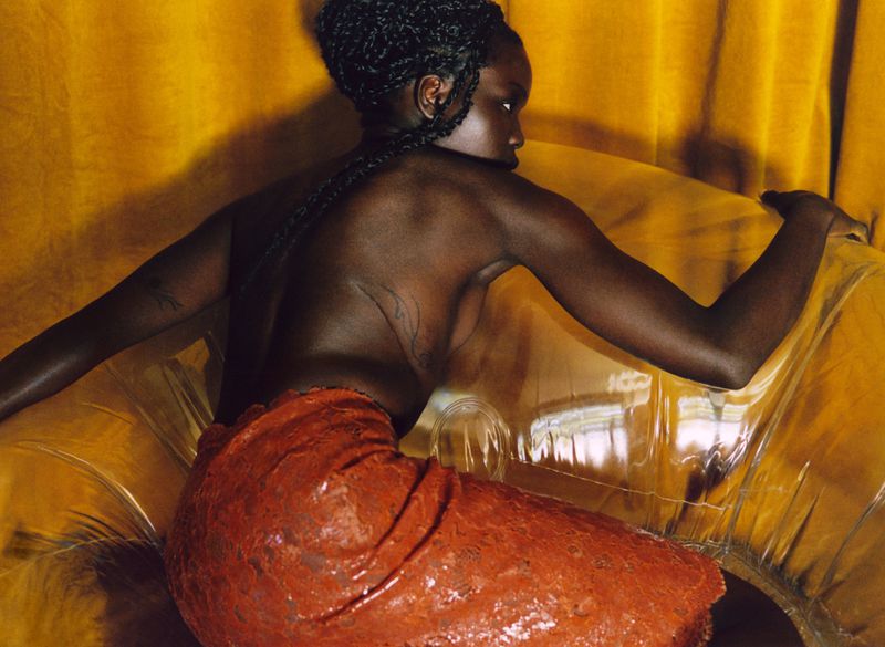Black woman with braided hair seen from behind posing on a transparent inflatable chair against yellow fabric