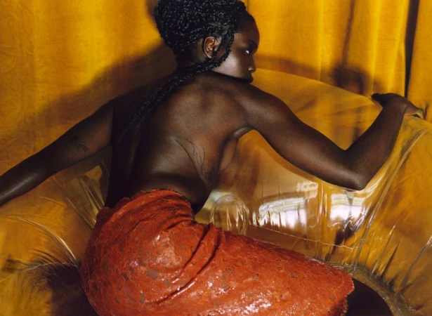 Black woman with braided hair seen from behind posing on a transparent inflatable chair against yellow fabric