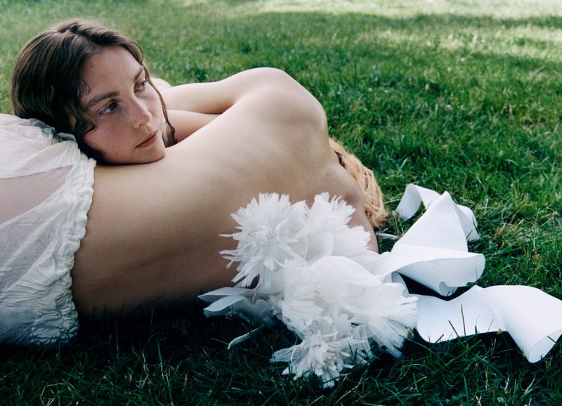 A young woman lying down on a green grass lawn with white decorative garments and ribbons near her.
