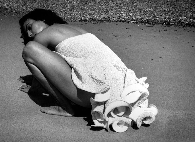 Model crouching on a sandy beach wearing a textured white garment with ruffled petal details for AnOther Magazine