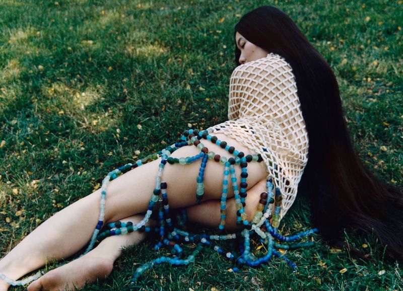 Woman with long dark hair wearing a mesh top and beaded skirt lying on green grass in AnOther Magazine editorial.