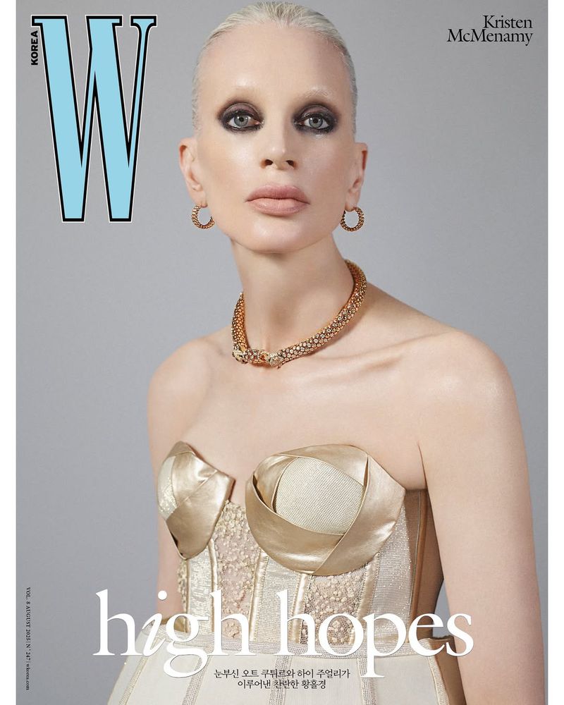 Magazine cover of Kristen McMenamy wearing jewelry and a strapless gold and white gown in a studio with a neutral backdrop.