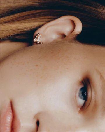 Extreme close-up portrait of a woman wearing a gold ear cuff with small embedded stones, highlighting freckles and natural skin texture.