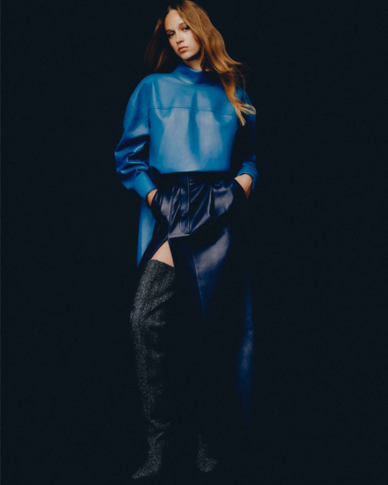 Model in a bright blue top and dark leather skirt with tall boots against a dark background for Mixte In the Shadows