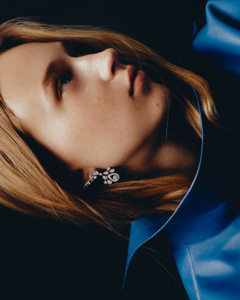 Close-up profile portrait of a woman wearing a blue garment and diamond earrings against a dark background