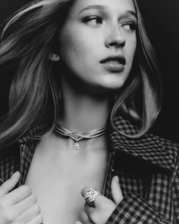 Close-up portrait of a woman wearing a diamond necklace, ring, and houndstooth jacket against a dark background.