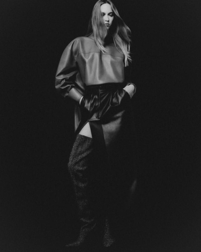 Model with long blonde hair wearing a dark leather top and skirt with thigh-high boots against a black backdrop