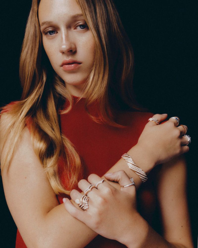 Close-up portrait of a woman wearing a red top and multiple diamond rings and a bracelet against a dark background.