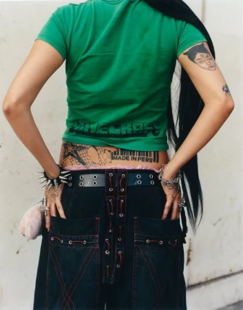 Rear view of a person wearing a green t-shirt, spiked bracelet, and black trousers with contrast lacing against a light wall