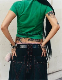 Rear view of a person wearing a green t-shirt, spiked bracelet, and black trousers with contrast lacing against a light wall