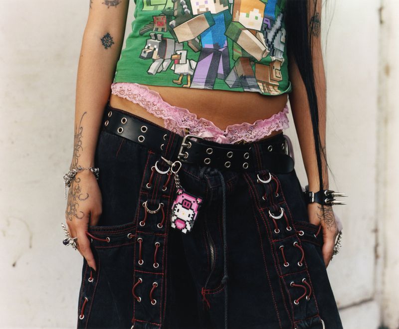 Close-up of a person wearing a green pixelated graphic top, pink lace thong, black grommet belt, and dark baggy cargo trousers.