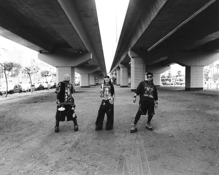 Three models in dark streetwear standing beneath a massive concrete highway overpass in an urban setting.