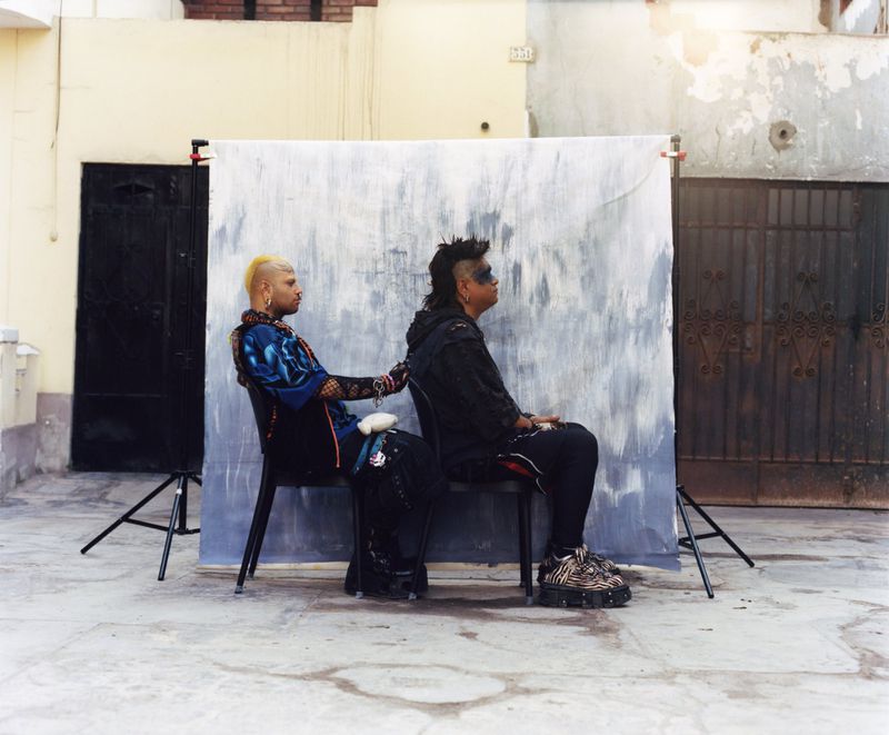 Two individuals with alternative styling seated back-to-back on chairs before a painted backdrop outdoors.