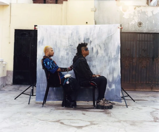 Two individuals with alternative styling seated back-to-back on chairs before a painted backdrop outdoors.