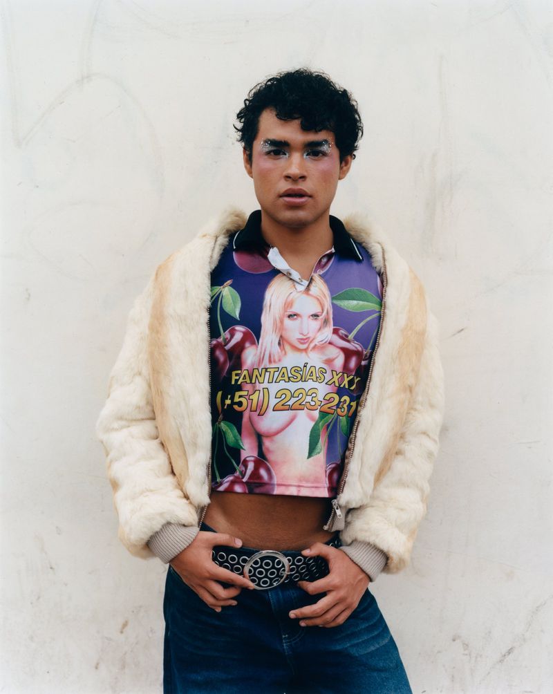 Young man wearing a graphic print crop top and faux fur jacket standing against a neutral wall for 72 Magazine.