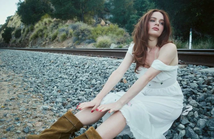 Actor Joey King seated on a gravel railroad track wearing a white dress and brown boots for Heroine Magazine