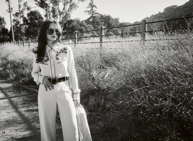 Monochrome portrait of actress Joey King wearing sunglasses and a patterned western shirt standing near a rural wooden fence.
