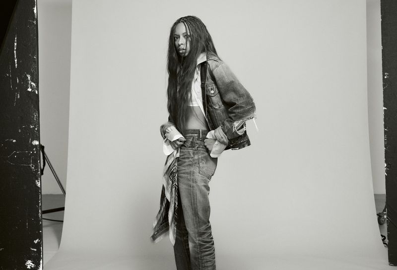 Black and white editorial portrait of Ravyn Lenae wearing denim jacket and jeans against a plain studio background.