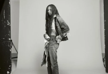 Black and white editorial portrait of Ravyn Lenae wearing denim jacket and jeans against a plain studio background.