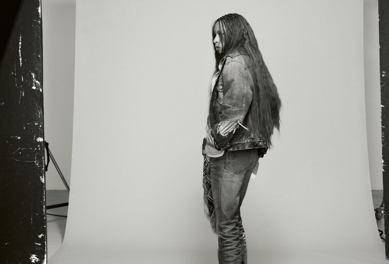 Black woman with long hair wearing a denim jacket and jeans standing against a grey studio seamless backdrop.