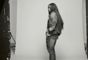Black woman with long hair wearing a denim jacket and jeans standing against a grey studio seamless backdrop.