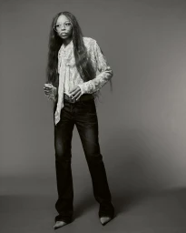 Portrait of Ravyn Lenae wearing a patterned blouse and dark trousers against a plain studio backdrop