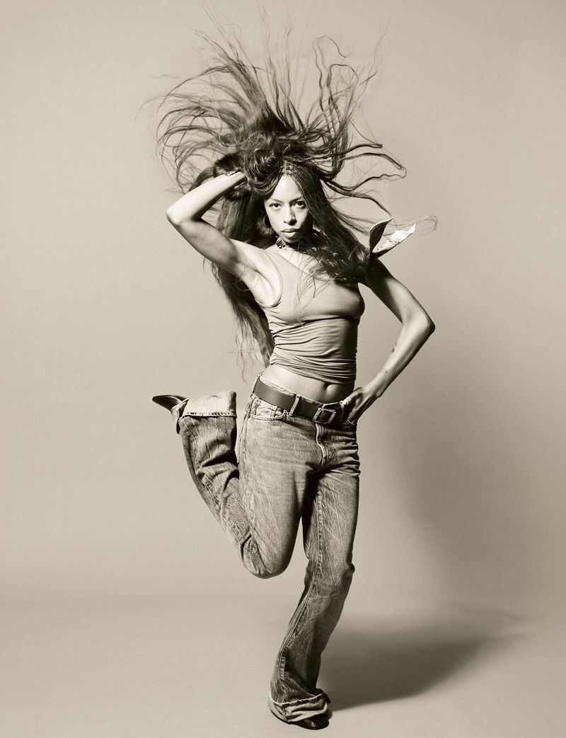 Ravyn Lenae jumping in mid-air with long dark hair blowing upward against a plain grey studio backdrop for Heroine Magazine.