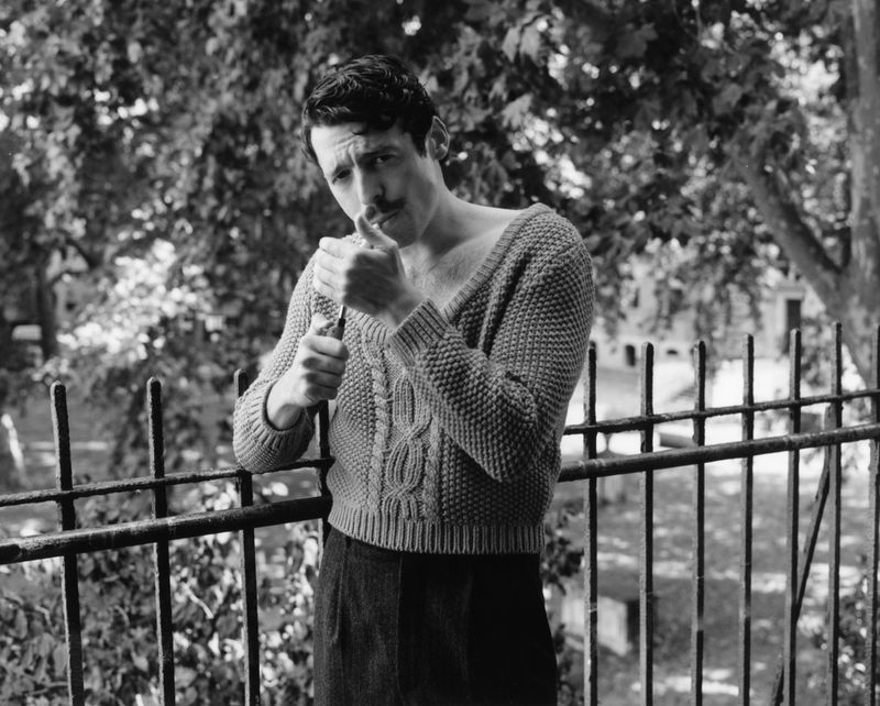 Anthony Boyle lighting a cigarette behind a metal fence wearing a cable-knit sweater in a black and white portrait.