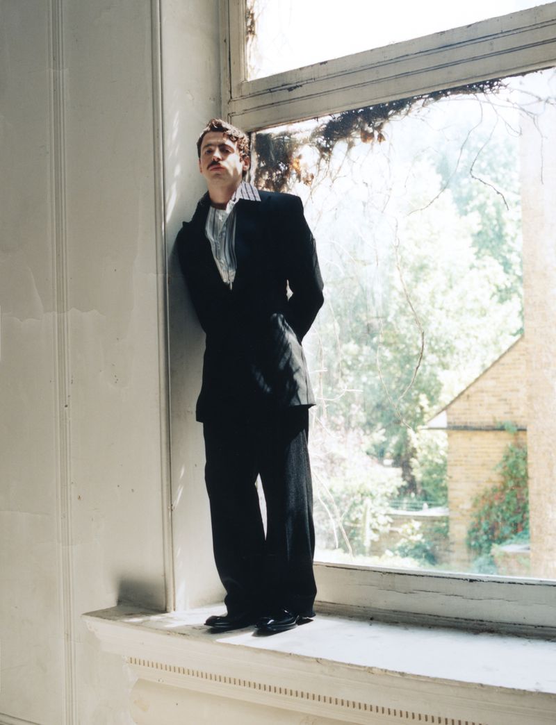 Anthony Boyle standing in a black suit leaning against a large window frame in an indoor setting.