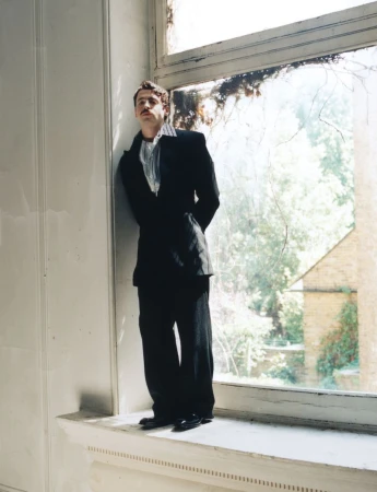 Anthony Boyle standing in a black suit leaning against a large window frame in an indoor setting.