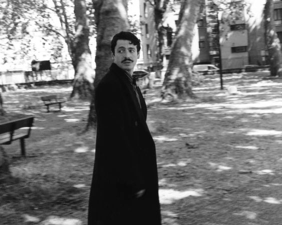 Anthony Boyle in a dark long coat walking in a park with large trees and benches in the background