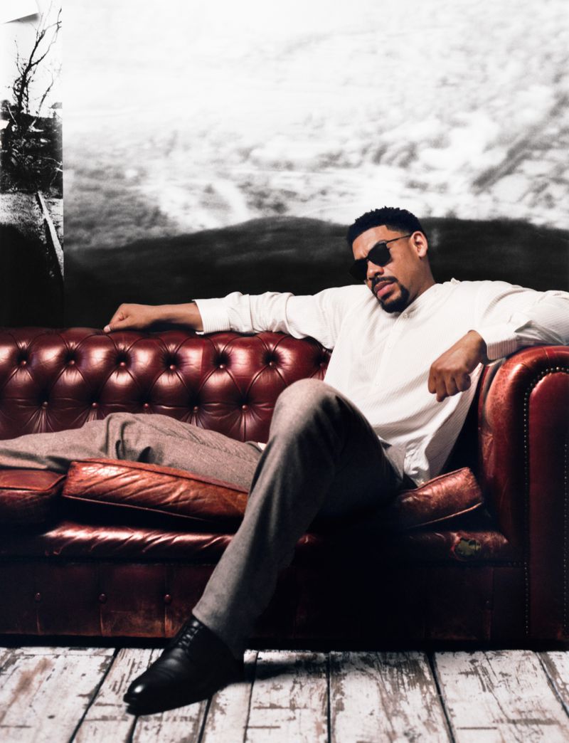 Actor Aaron Pierre seated on a red leather chesterfield sofa wearing a white shirt and sunglasses against a monochrome landscape backdrop.
