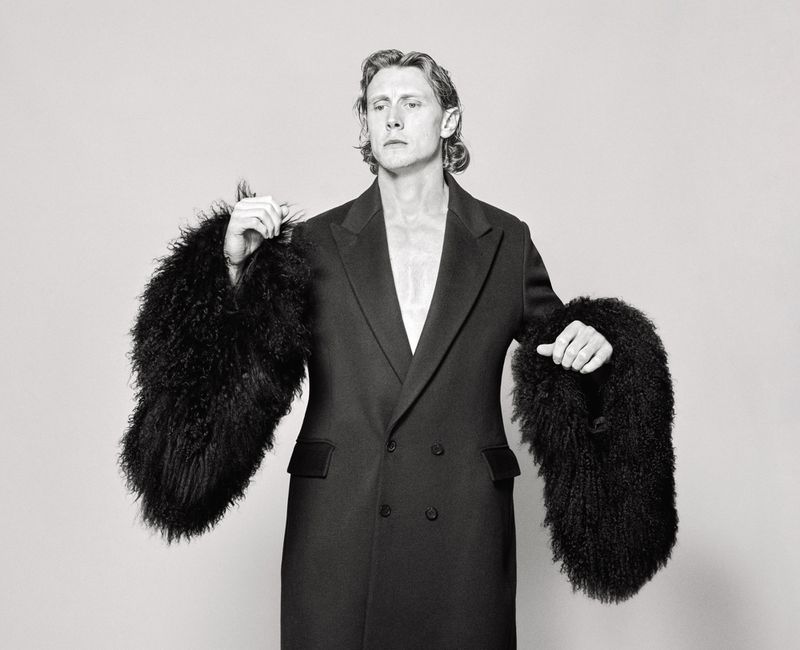 George MacKay wearing a black coat with furry sleeves against a light seamless background for HERO Magazine.