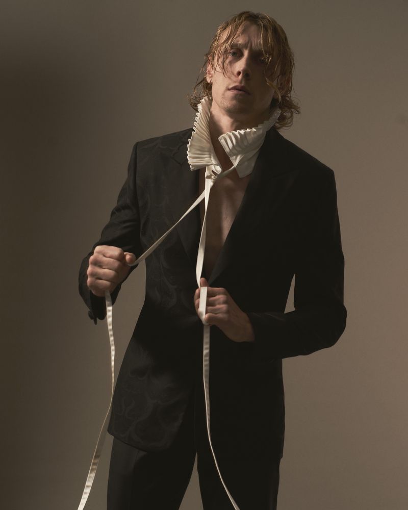 Portrait of actor George MacKay wearing a dark jacket with a tall pleated white collar for HERO Magazine